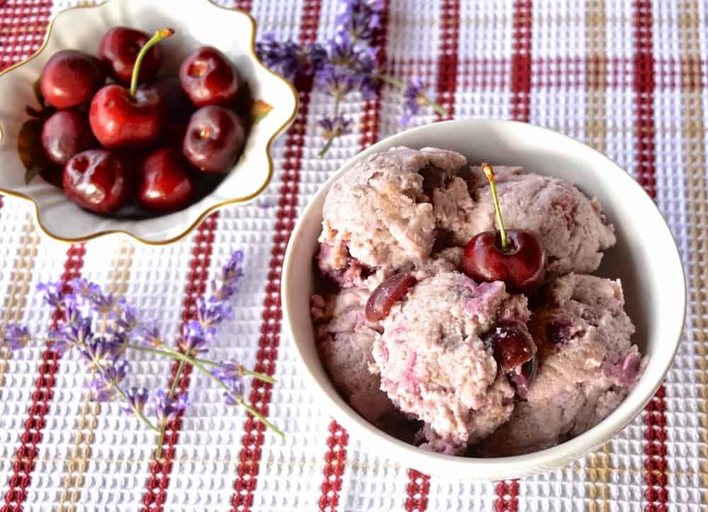 A bowl of cherry ice cream topped with a fresh cherry with lavender flowers around the bowl.