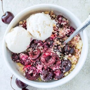 A bowl of paleo cherry crisp with two scoops of vanilla ice cream on it and a spoon in the bowl.