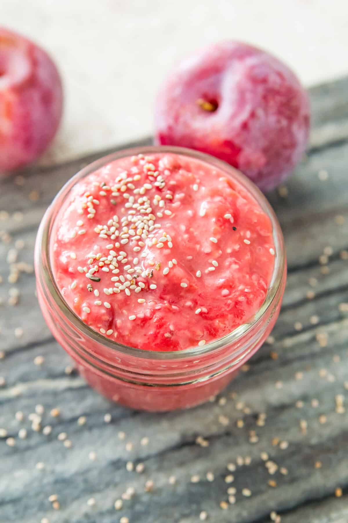 A jar of plum chia jam with chia seeds on top of it and two plums beside the jar. 