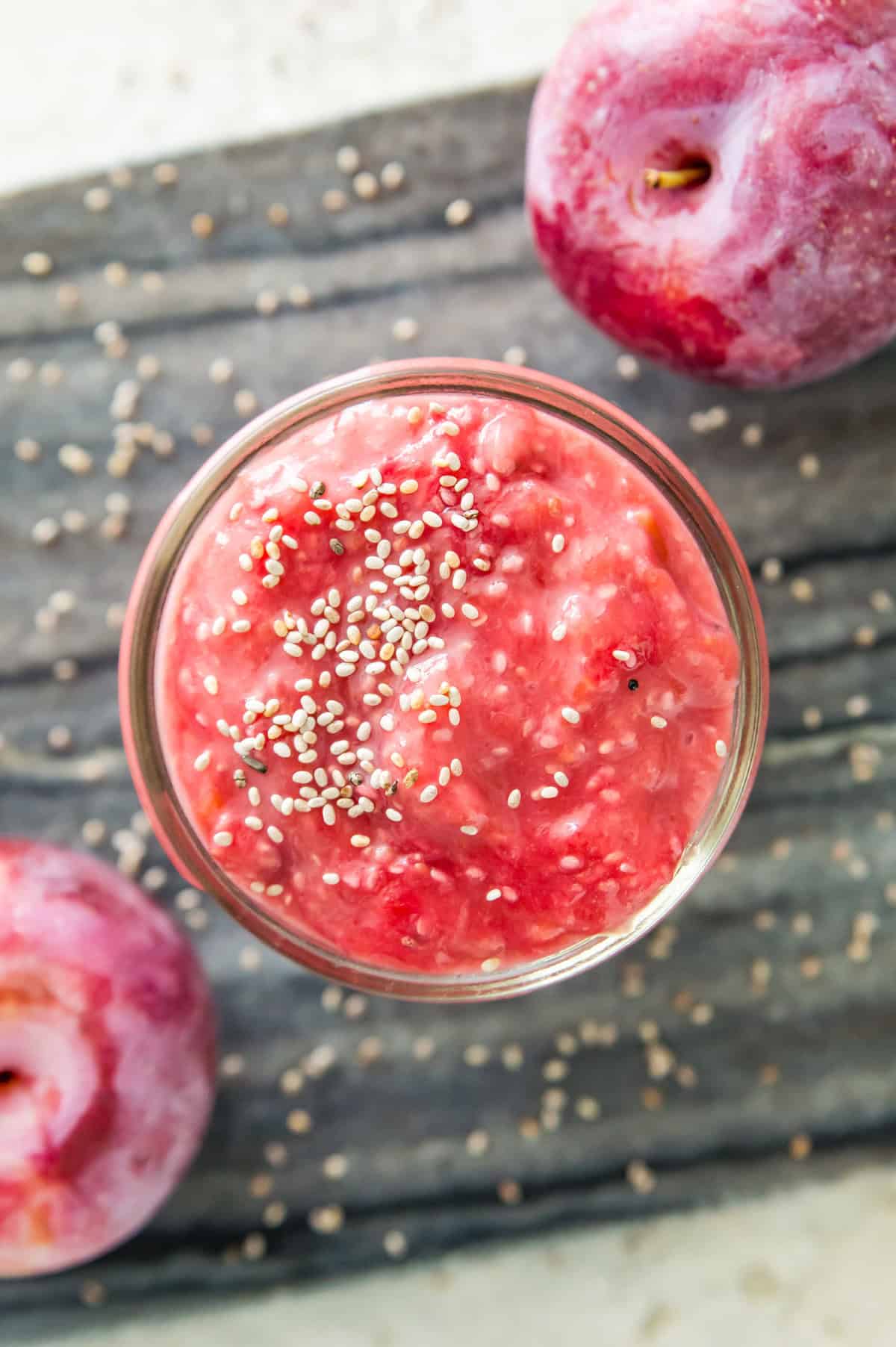 A small jar filled with plum jam with chia seeds on top of it and two plums beside the jar.