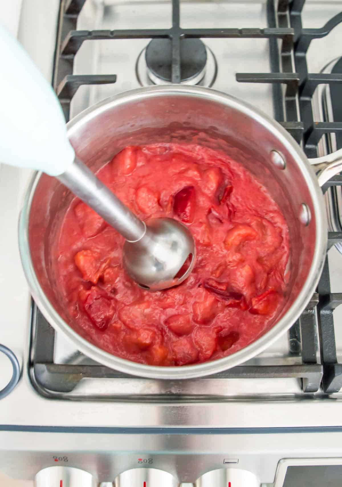 A pot on the stovetop with cooked plums in it and an immersion blender blending the plums. 