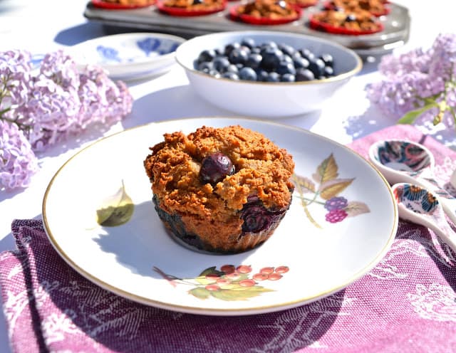 A paleo coconut blueberry muffin on a plate with a bowl of blueberries behind it.
