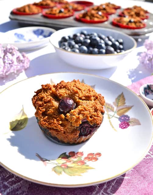 A paleo coconut blueberry muffin on a plate with a bowl of blueberries behind it.