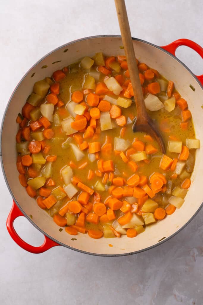 A pot filled with chopped carrots, turnips and onions cooking on a stovetop with a spoon in the pot.