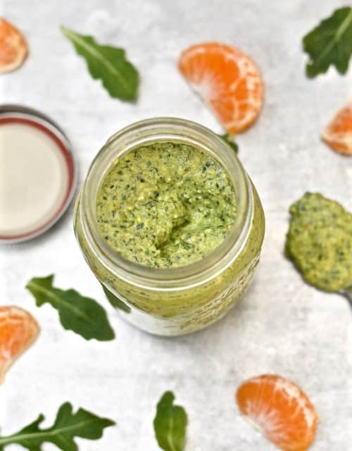 A jar of pesto with orange pieces and arugula leaves around it.