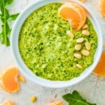 A bowl filled with arugula pesto garnished with pine nuts and an orange slice.