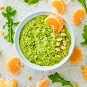 A white bowl filled with arugula pesto and garnished with pine nuts and an orange slice.