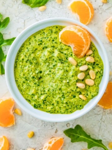A white bowl filled with arugula pesto and garnished with pine nuts and an orange slice.