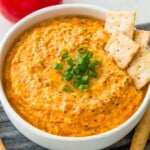 A bowl of vegan roasted red pepper dip garnished with chopped green onion and with four crackers in it.