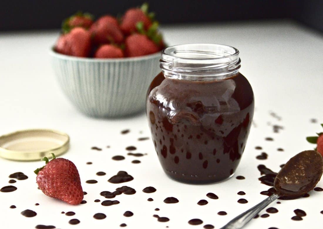 A jar filled with a healthy chocolate sauce with a bowl of strawberries behind it.
