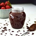 A jar of chocolate sauce with a bowl of strawberries behind it.