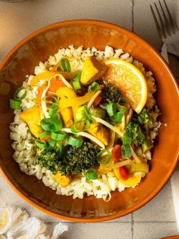 A bowl of orange ginger chicken stir fry over cauliflower rice with a fork next to the bowl.