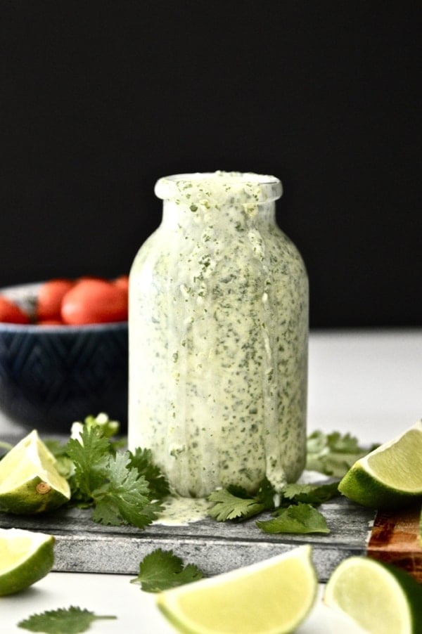 A bottle of vegan cilantro lime dressing surround by cilantro and lime wedges. 