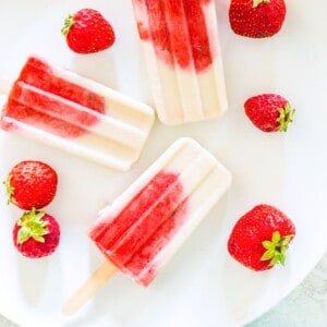 Three strawberry vanilla popsicles on a white plate surrounded by fresh strawberries.