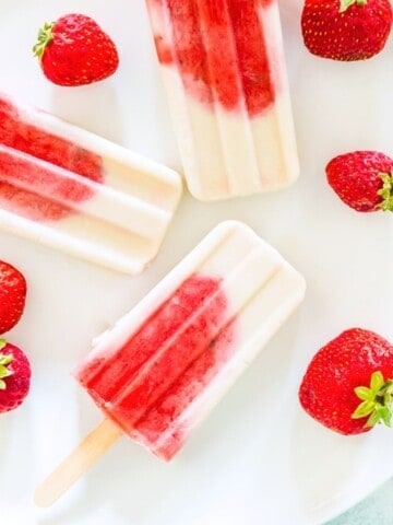 Three strawberry vanilla popsicles on a white plate surrounded by fresh strawberries.