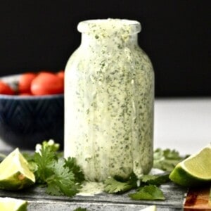 A bottle of vegan cilantro lime dressing surrounded by fresh cilantro and lime wedges.