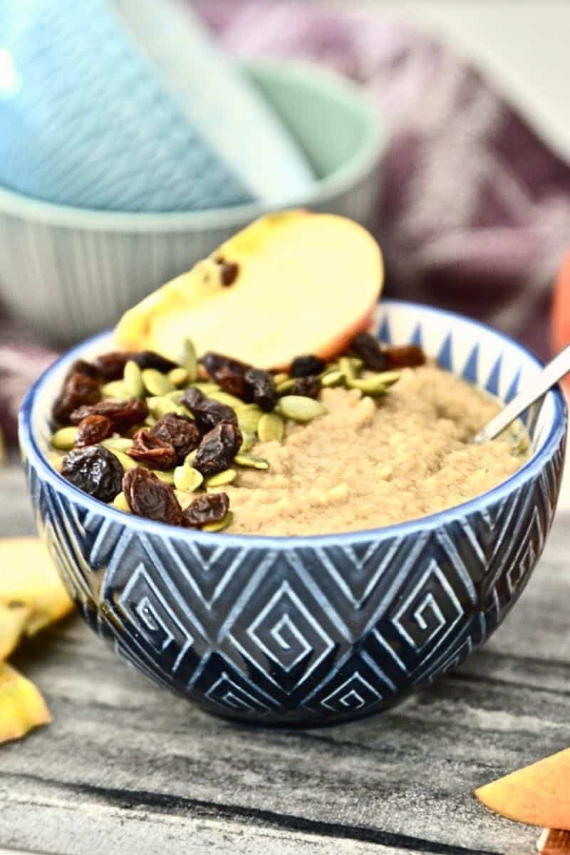 A bowl of oatmeal topped with raisins, pumpkin seeds and a slice of apple.