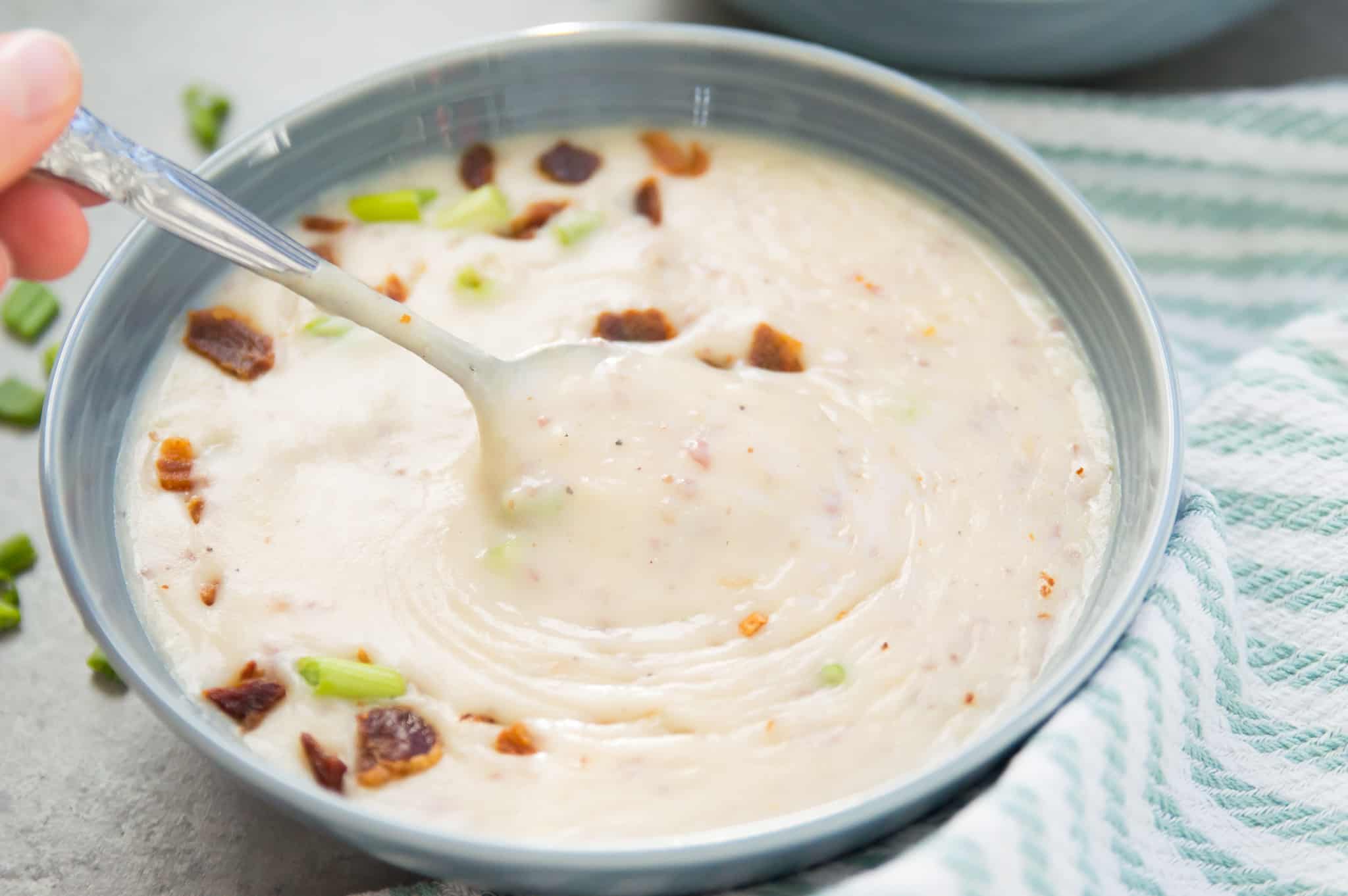 A bowl of creamy potato soup topped with bacon pieces and chives with a spoon being dipped into it.