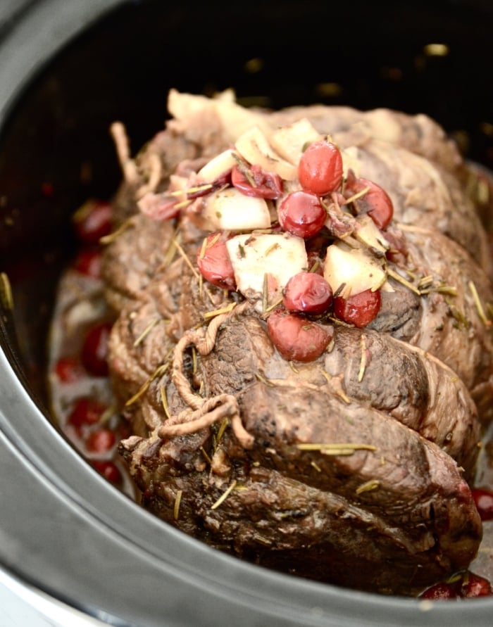 A beef roast in a slow cooker with cranberries and onions  on top of it.