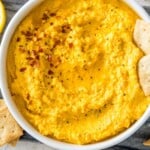 A bowl of carrot dip topped with chili flakes and with crackers in it.