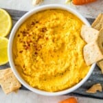A bowl of carrot dip topped with chili flakes and two crackers.