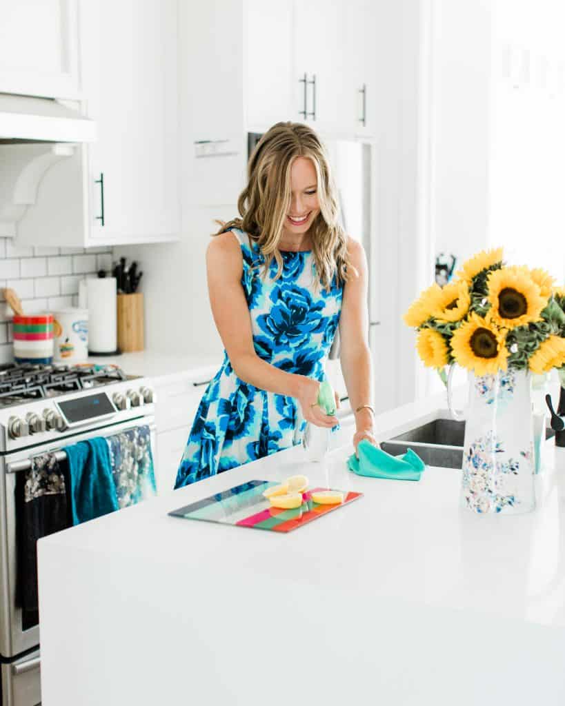 A girl with blond hair wearing a blue floral dress cleaning a kitchen counter.