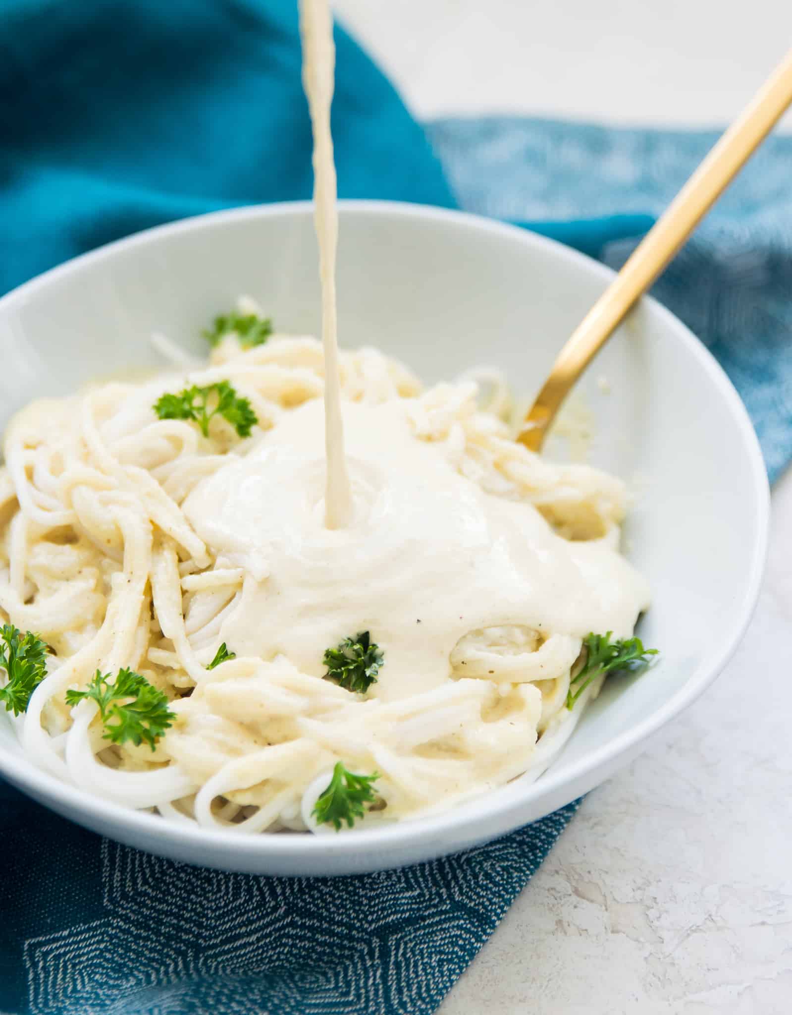 Dairy free Alfredo sauce being poured onto cooked pasta garnished with fresh parsley. 