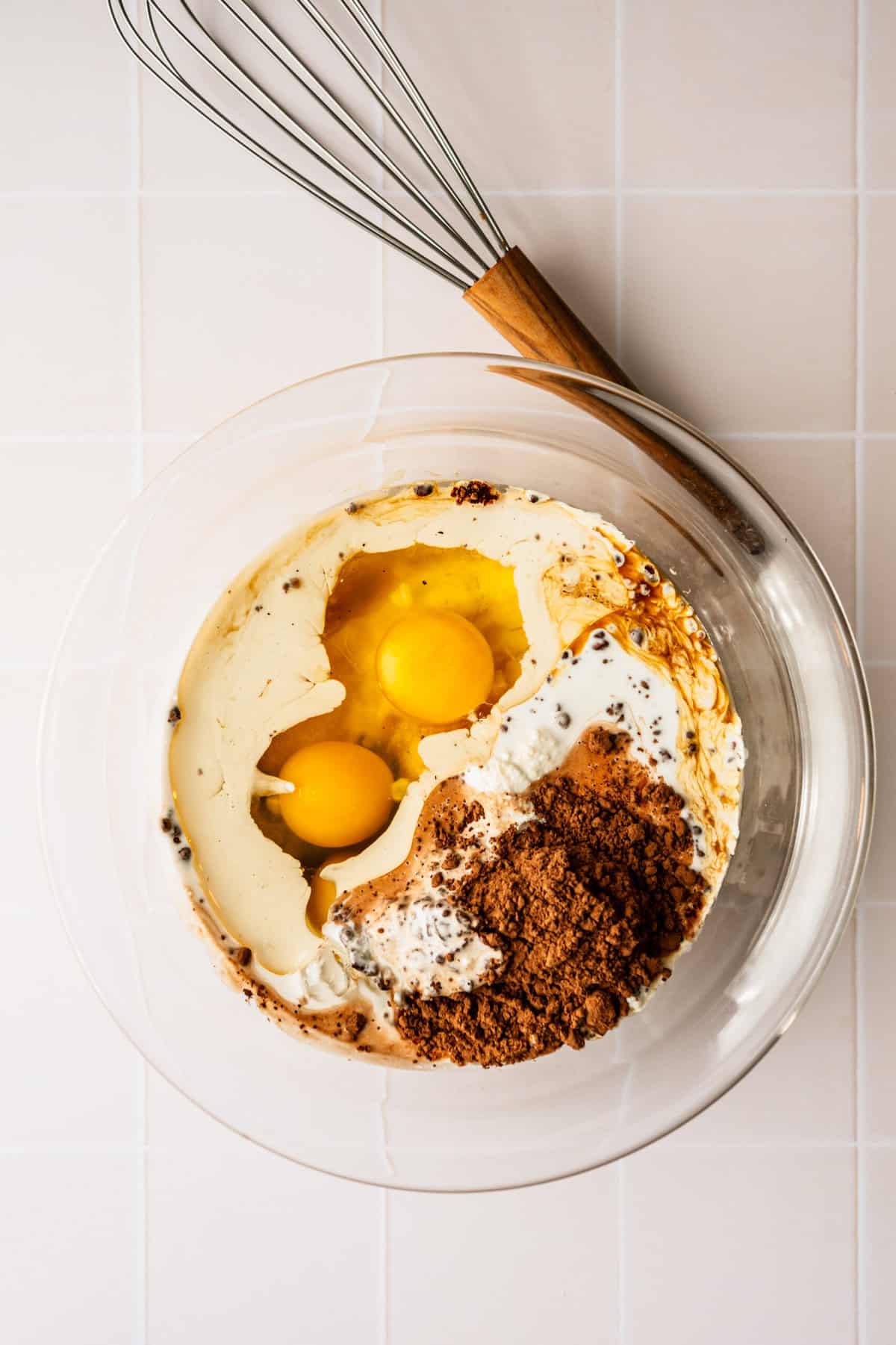 A glass bowl with eggs, cocoa powder and milk in it.