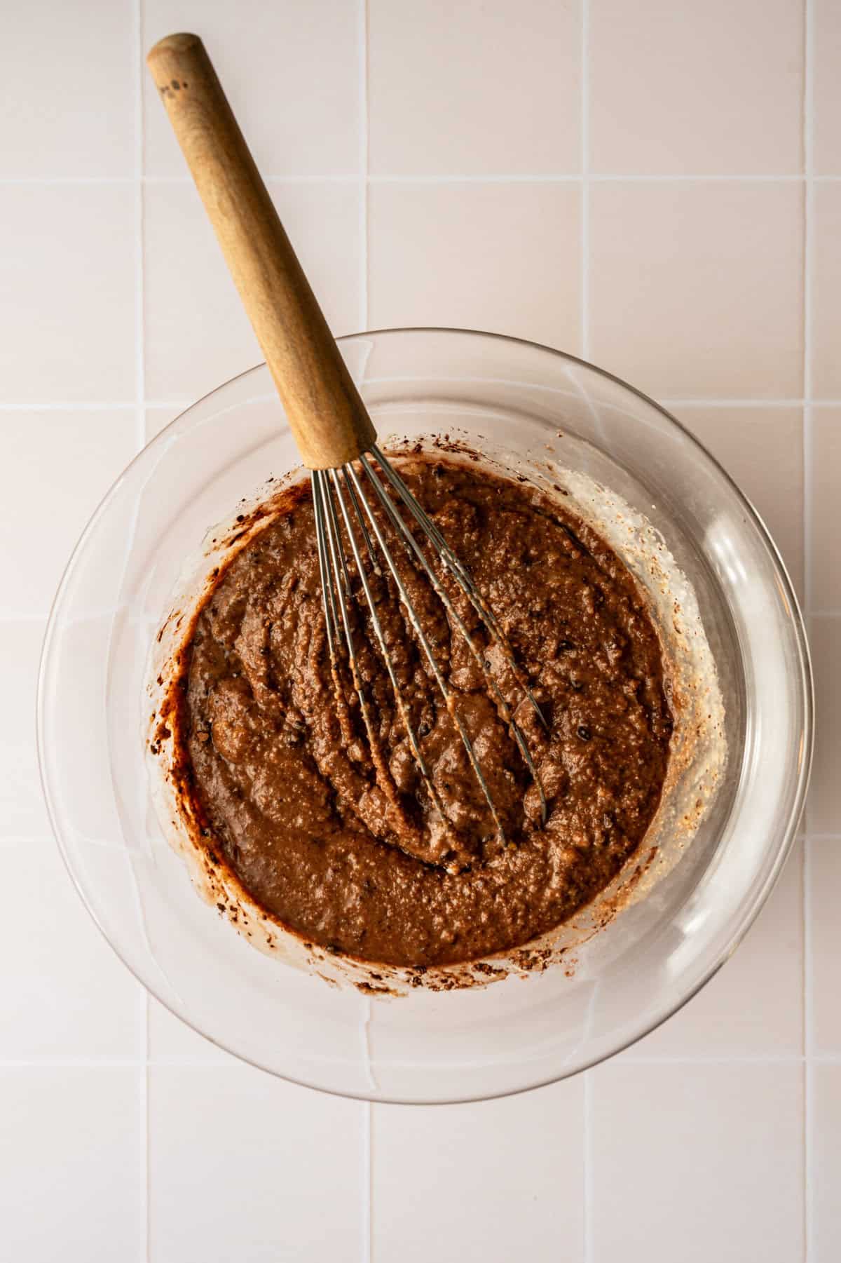 A glass bowl filled with a chocolate batter with a whisk in the batter.