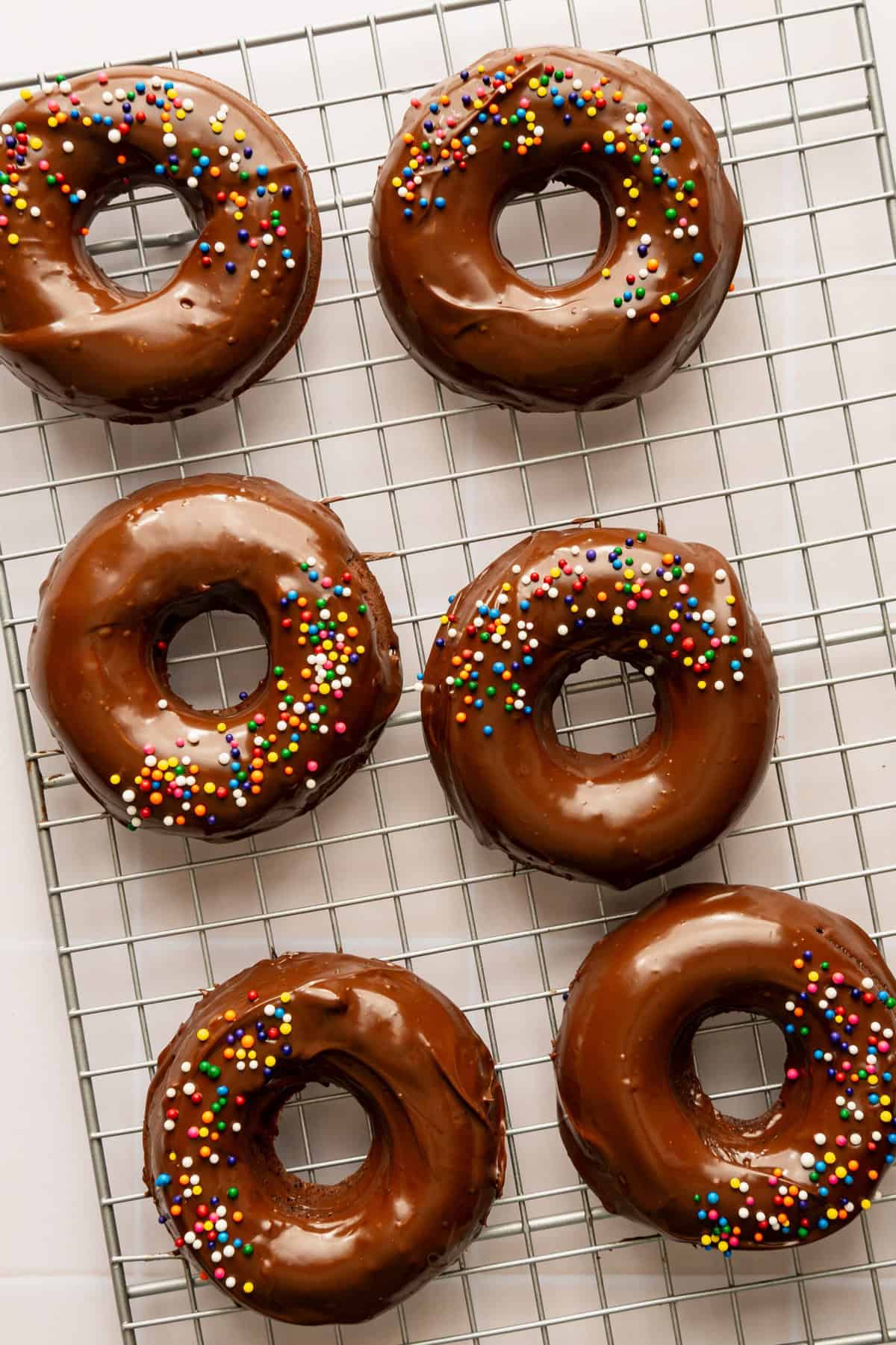 A batch of chocolate donuts topped with rainbow sprinkles on a wire baking rack.