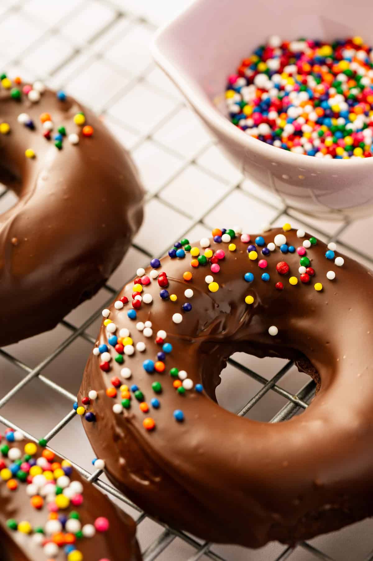 Chocolate donuts topped with rainbow sprinkles on a wire baking rack with a bowl of sprinkles next to them.