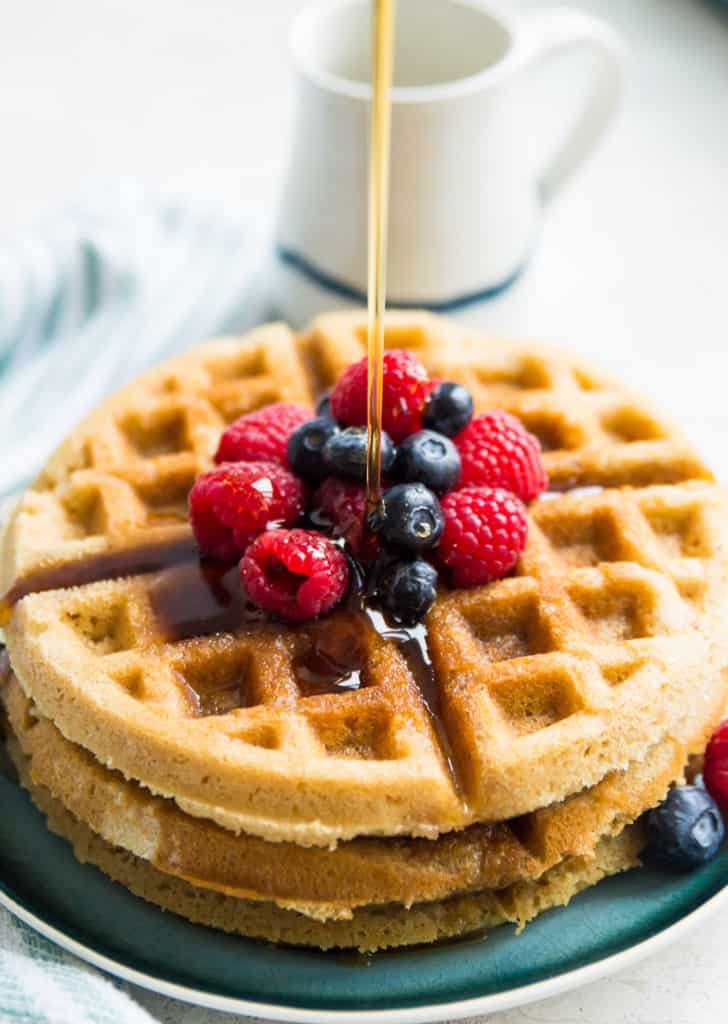 Paleo waffles topped with blueberries and raspberries with maple syrup being poured on them
