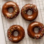 A batch of chocolate donuts topped with rainbow sprinkles on a wire baking rack.