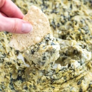 A chip being dipped into a bowl of vegan spinach artichoke dip.