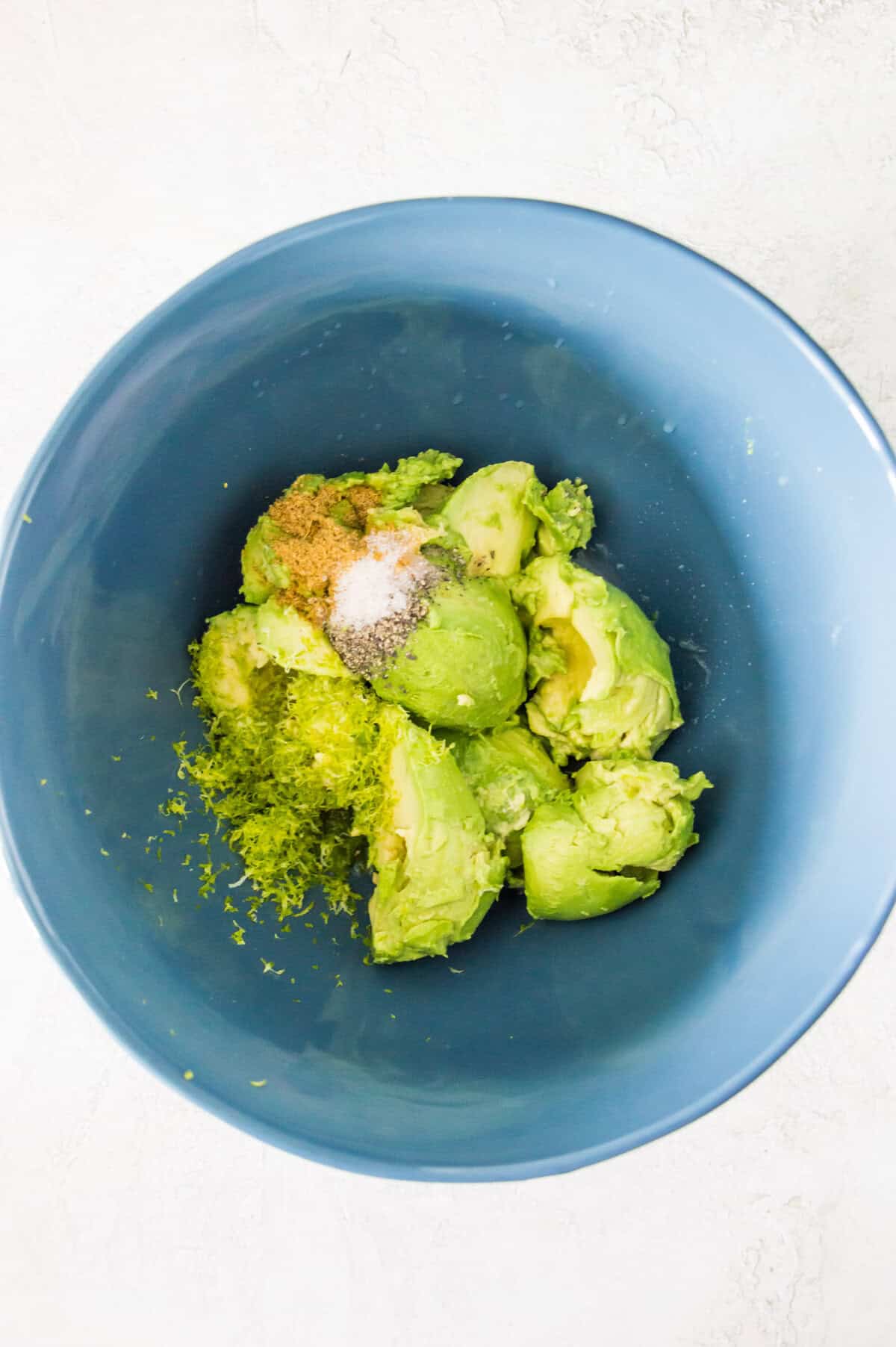 A bowl with avocado flesh, salt, ground black pepper, and cumin powder in it.