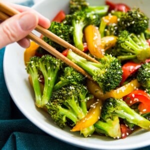 A bowl of cooked teriyaki broccoli with sesame seeds on it for garnish and chop sticks picking up a piece of broccoli.