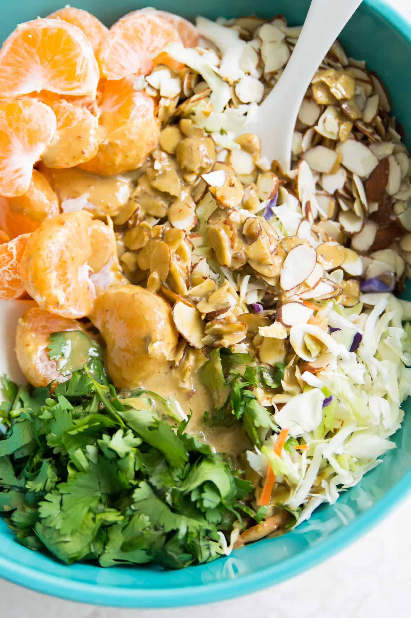 A bowl of coleslaw with mandarin oranges, almonds, cilantro and dressing on it.