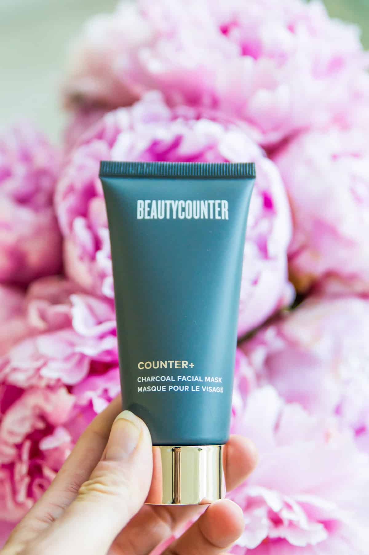 Beautycounter's charcoal mask being held over a batch of pink peonies. 