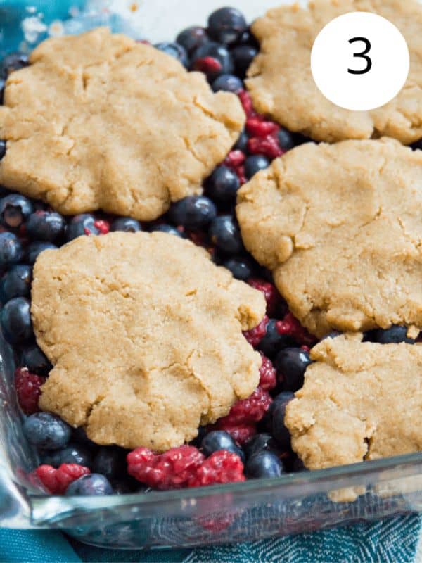 A glass baking dish with blueberries and raspberries in it topped with uncooked pieces of dough.