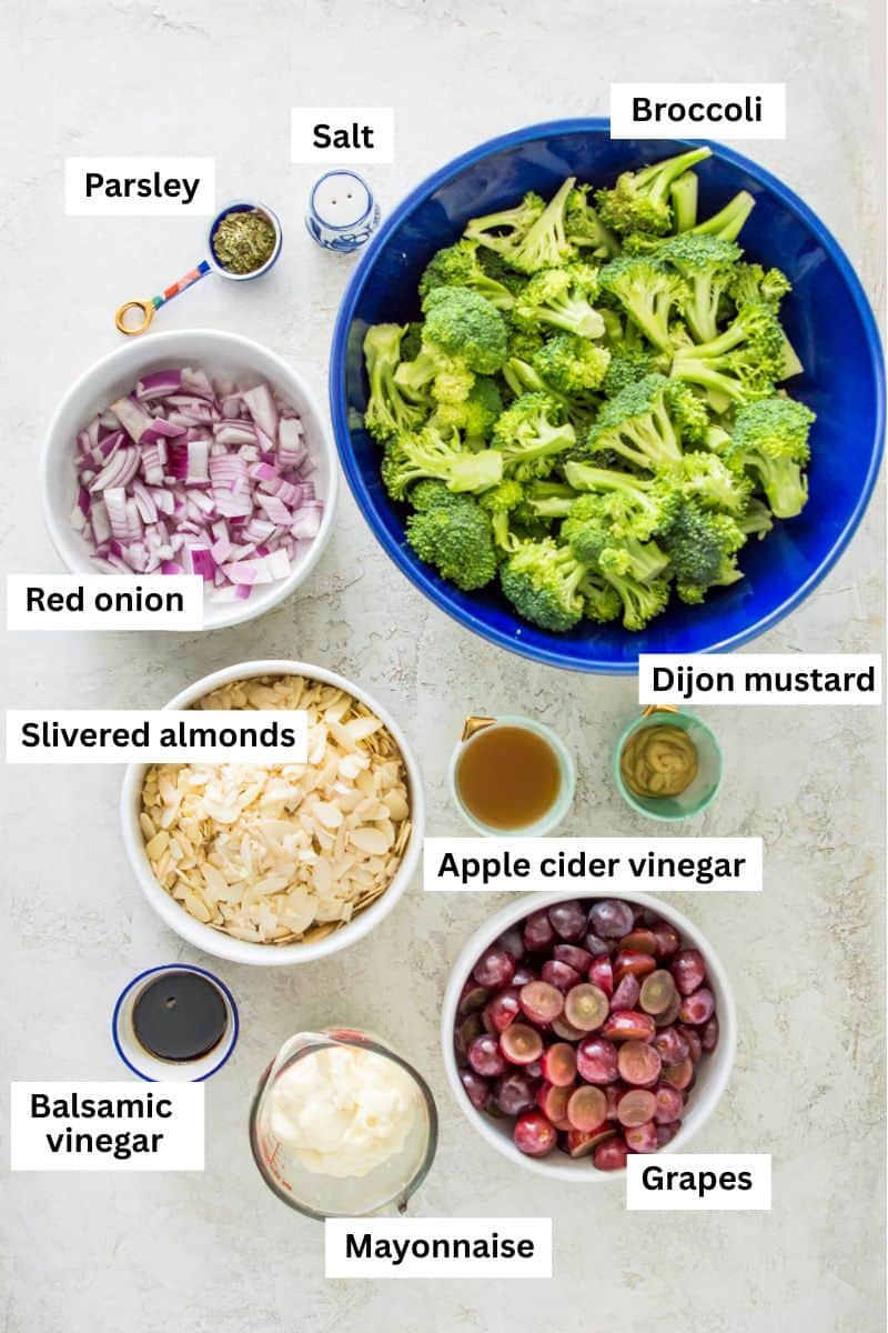 Foods in bowls including broccoli florets, chopped red onion, chopped red grapes, mayonnaise, salt, slivered almonds, parsley and vinegar. 