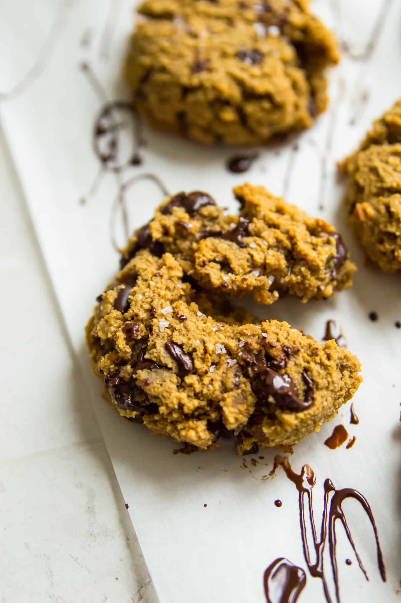 A pumpkin chocolate chip cookie on parchment paper drizzled in dark chocolate with coarse sea salt on top of it.