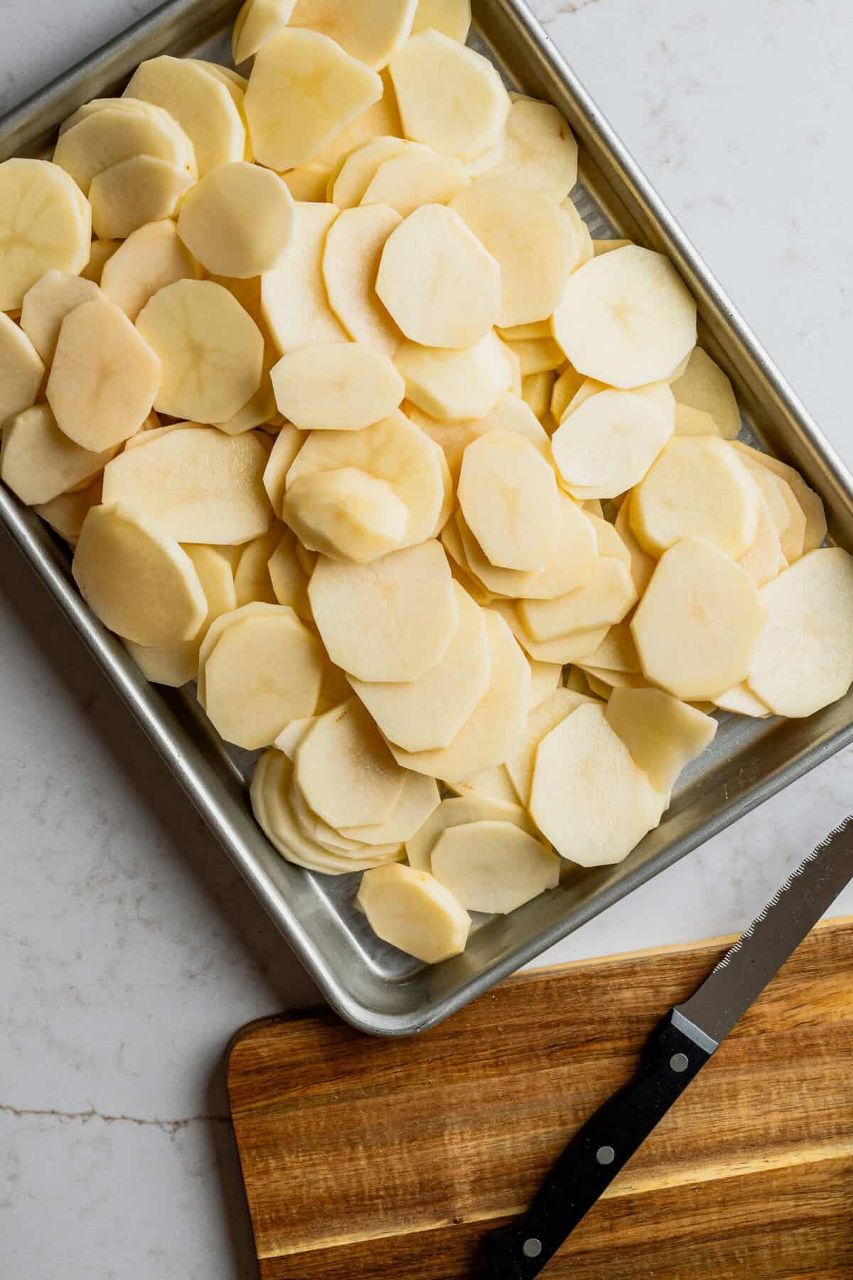 Thinly sliced potatoes in a metal pan with a knife and cutting board next to it.