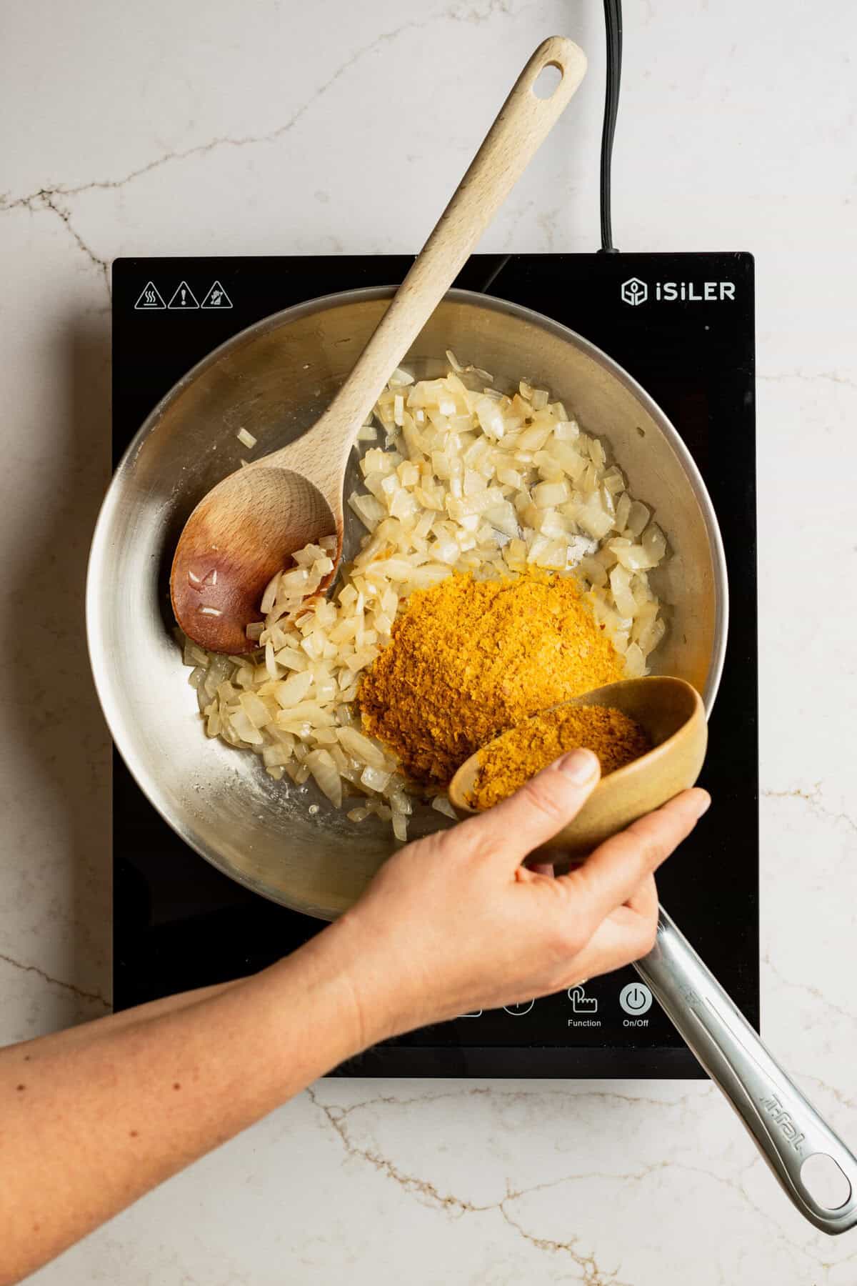 Chopped onions cooking in a pan with a wooden spoon in it and nutritional yeast being added to the pan. 
