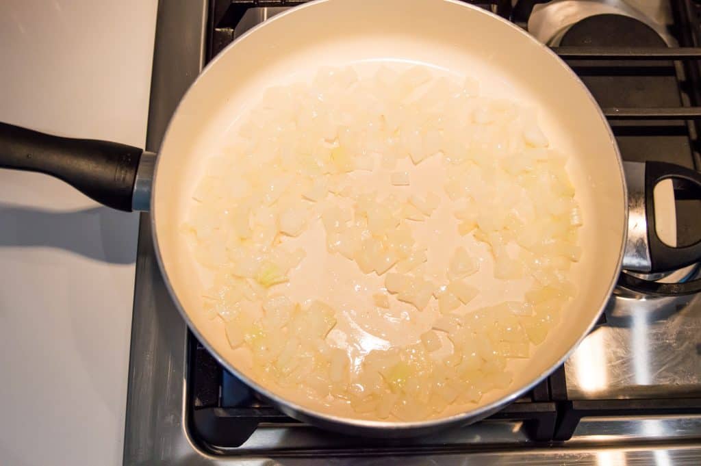 A large pan on the stovetop with chopped onions cooking in it.