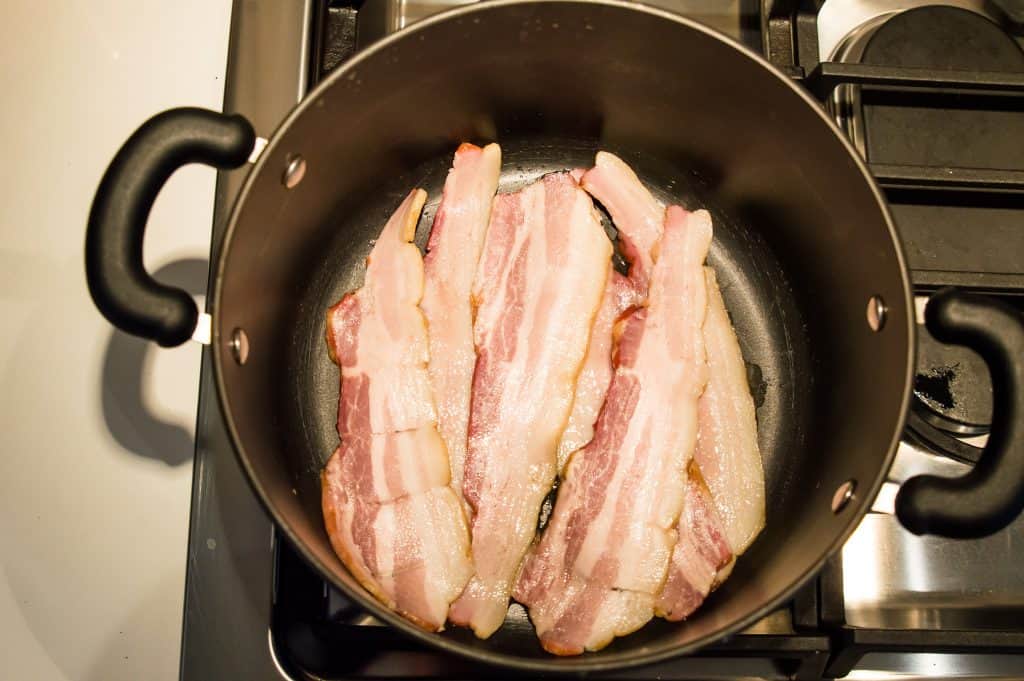 A pot on the stovetop with bacon being cooked in it.