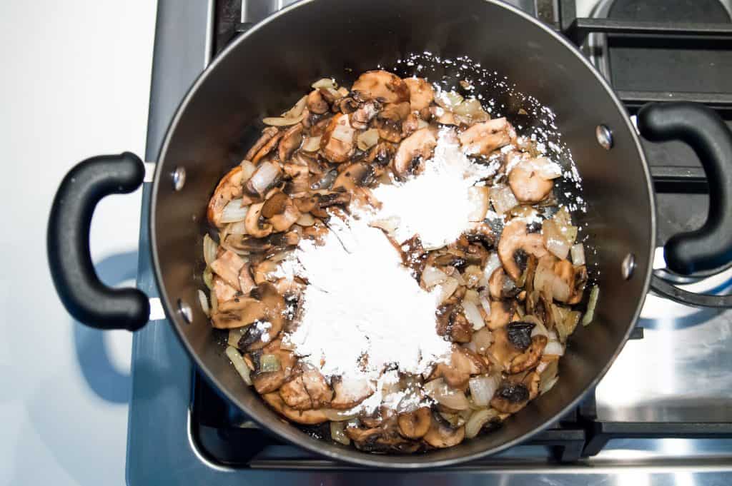 A large pot on the stove top with cooked mushrooms and cornstarch in it.
