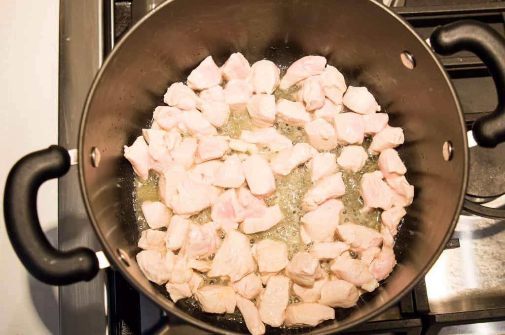 A black pot on the stovetop with chicken pieces cooking in it.