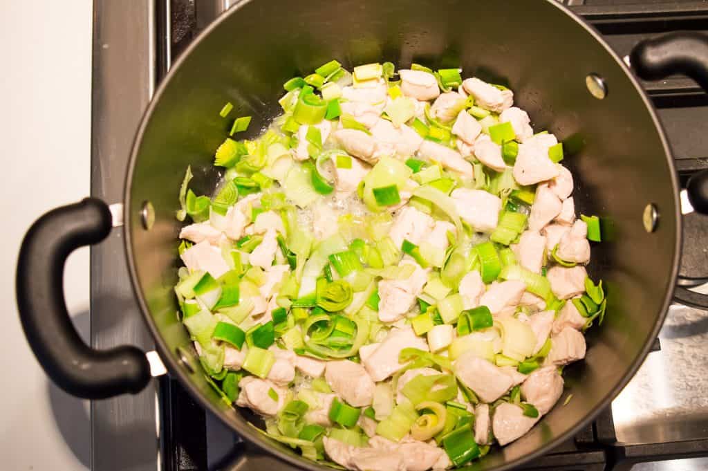 A large pot on the stove with cut chicken, leeks and garlic cooking in it.