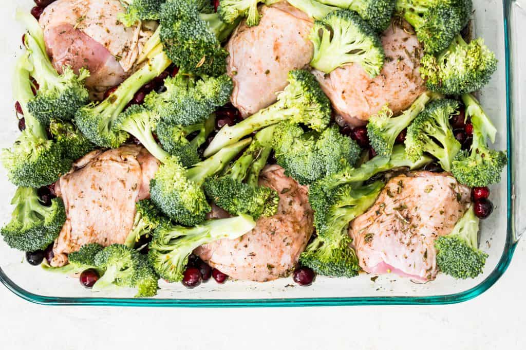 A baking dish with chicken thighs, chopped broccoli and fresh cranberries in it.