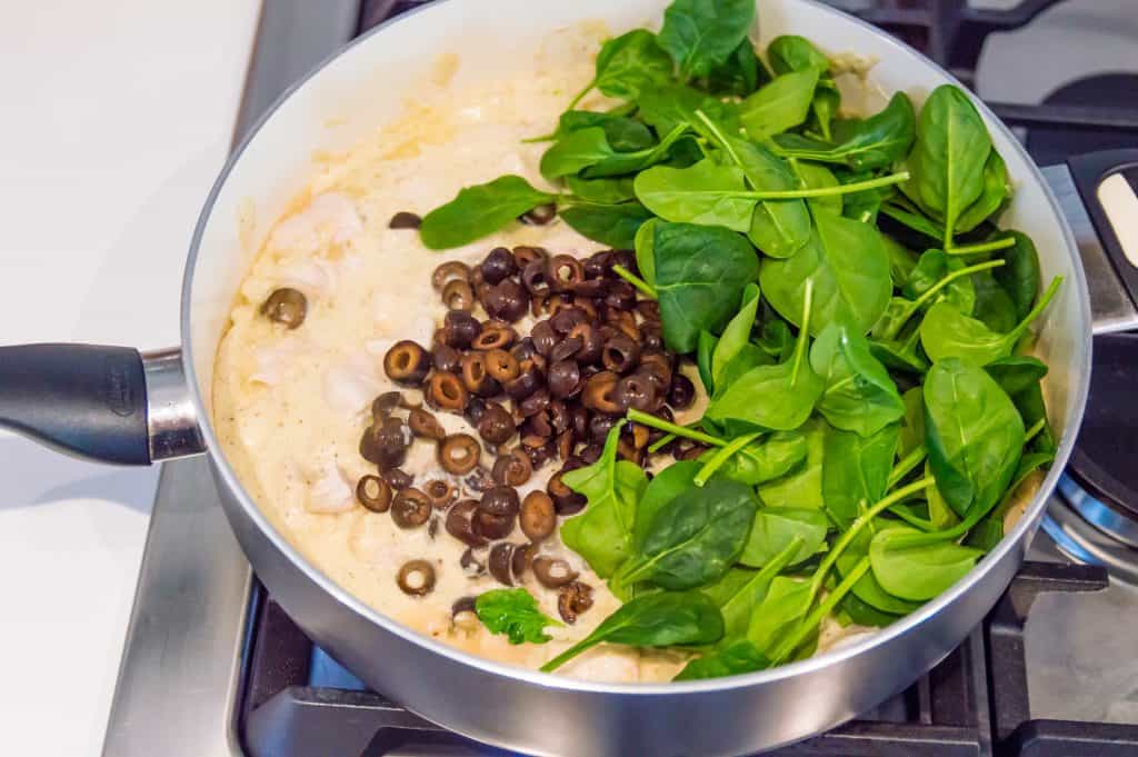 A pan with Alfredo Sauce, chicken pieces, fresh spinach and black olives in it on the stovetop.